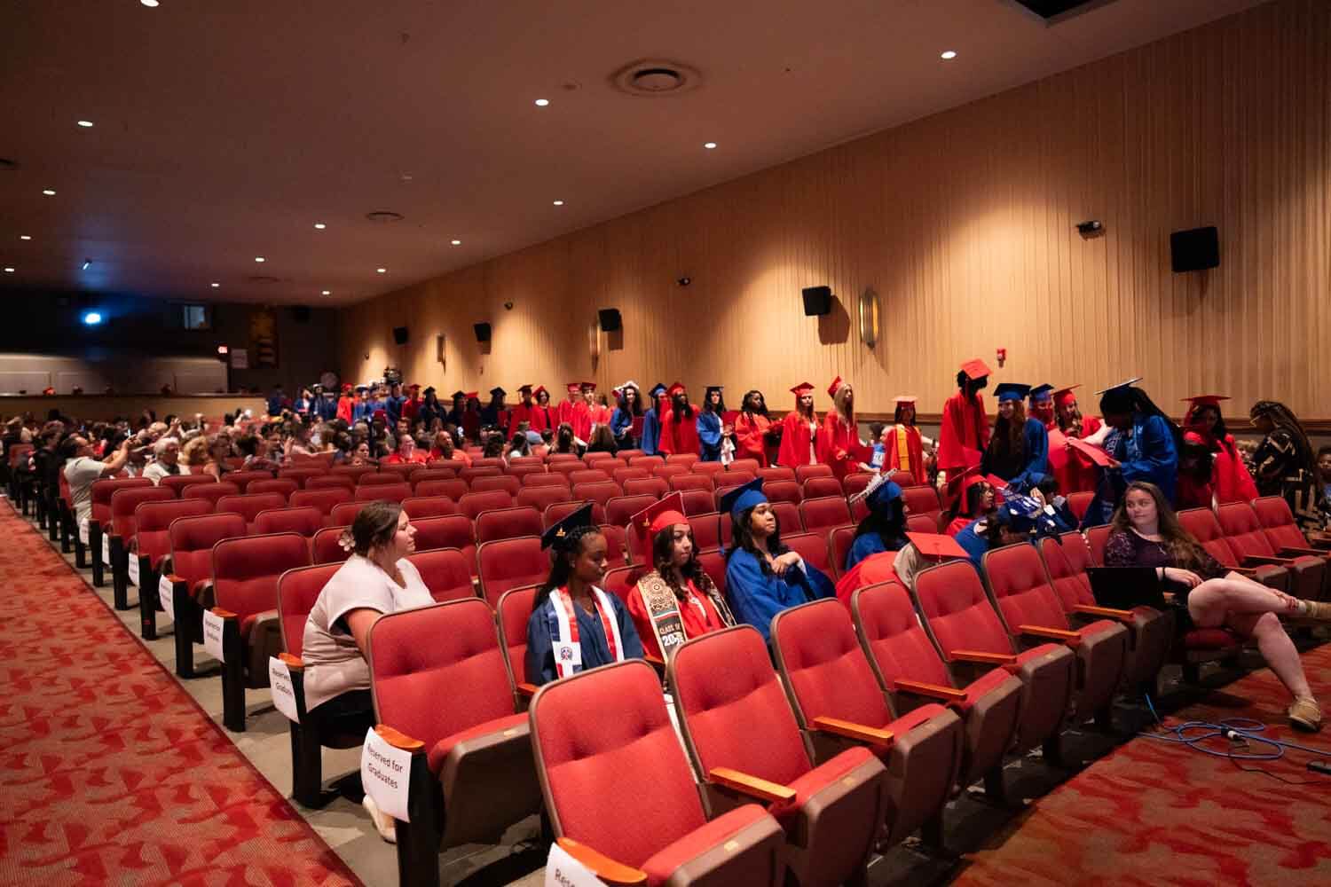 Ardayda qalin jebisay oo xidhan gownyo casaan iyo buluug ah iyo koofiyadaha ayaa buuxiya kuraasta fadhiga, qaar taagan oo sheekaysanaya.