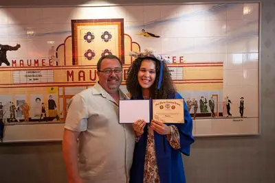 Gabadh yar oo dhoola cadeynaysa oo huwan shaadhka qalinjebinta buluuga ah iyo koofiyadda ayaa sita shahaado, iyadoo taagan nin ag taagan.